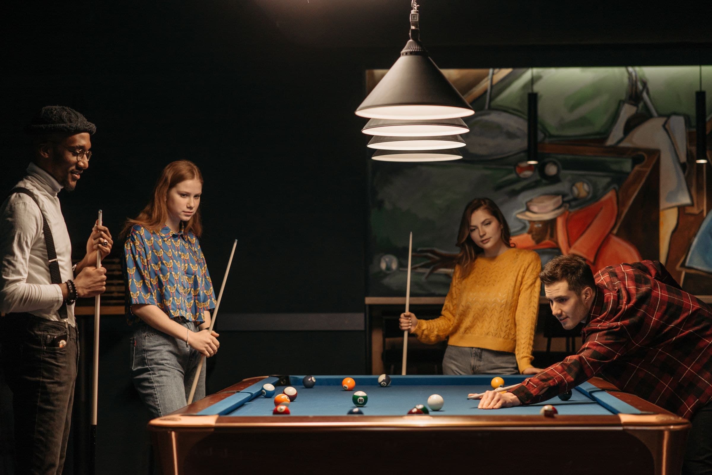 Friends gathered around a pool table in a warmly lit venue as one player lines up a shot